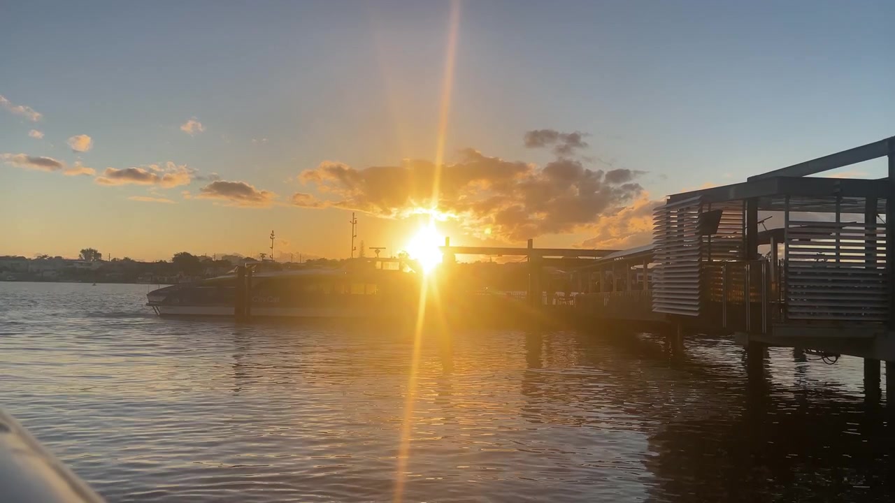 Um pôr do sol laranja brilhante ilumina a orla do terminal de balsas, criando um reflexo dourado vibrante sobre a água circundante