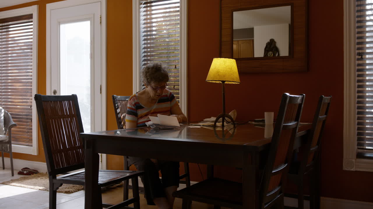 An elderly mother sits alone at a dining room table, sorting through a pile of bills, and reviewing paperwork. Warmly lit, cozy home, by a table lamp. Orange painted walls, and brown furniture. 24fps