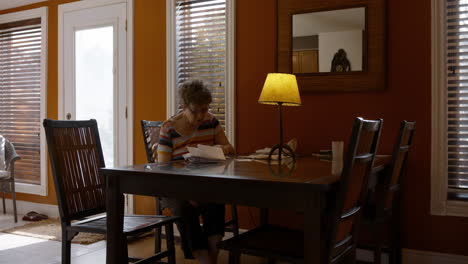 an elderly mother sits alone at a dining room table, sorting through a pile of bills, and reviewing paperwork. warmly lit, cozy home, by a table lamp. orange painted walls, and brown furniture. 24fps