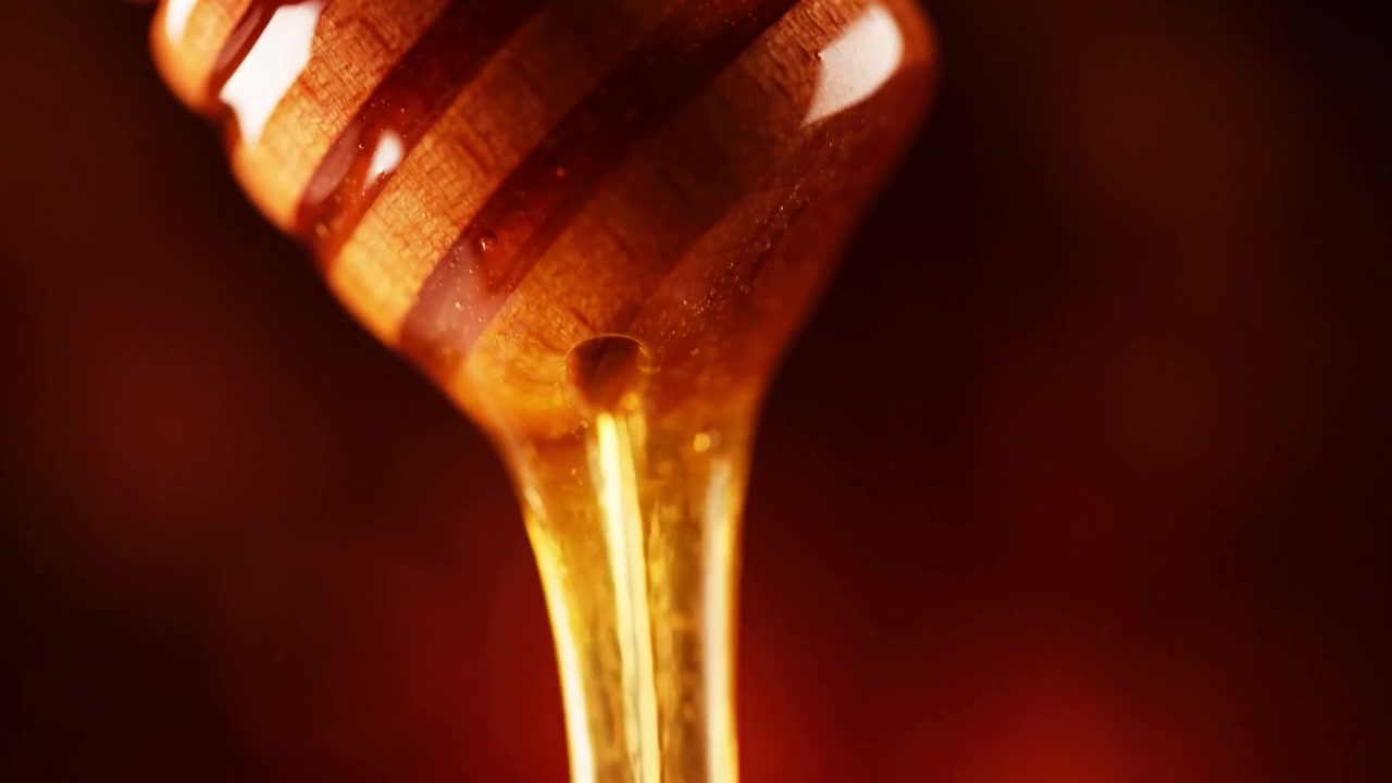 Macro primo piano di miele dorato che cola da un mestolo di legno su sfondo scuro