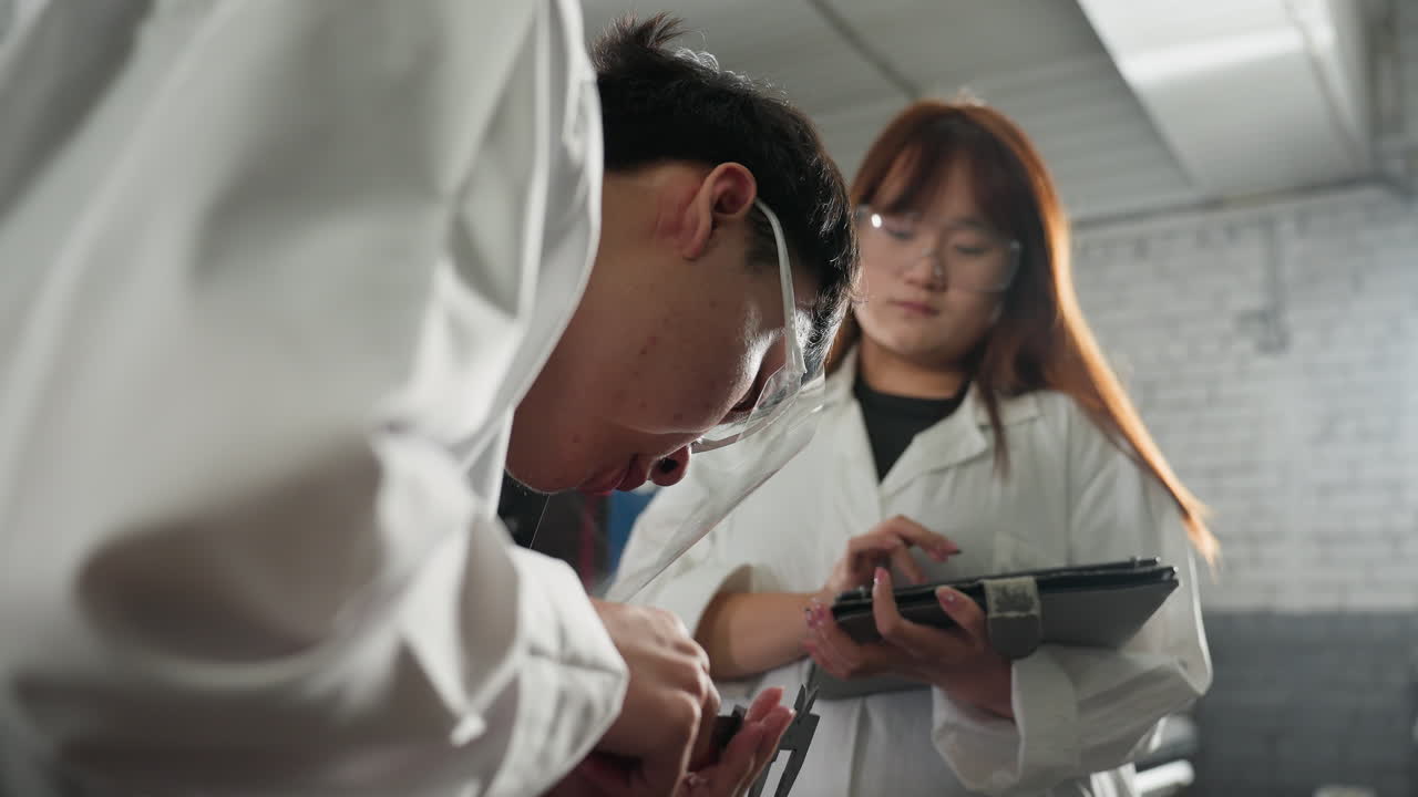 보호 안경을 착용한 기계 작업실의 엔지니어, 베르니에 칼리퍼로 엔진 크기를 측정, 근처의 여성 동료 운영 태블릿, 데이터 기록