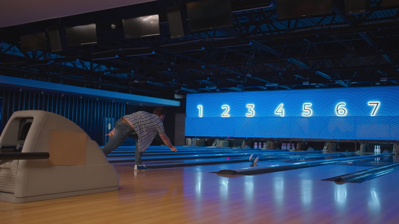 In a plaid shirt a man in a bowling alley throws a ball in slow motion and knocks down skittles. Play bowling. Throw balls on the floor of the bowling club
