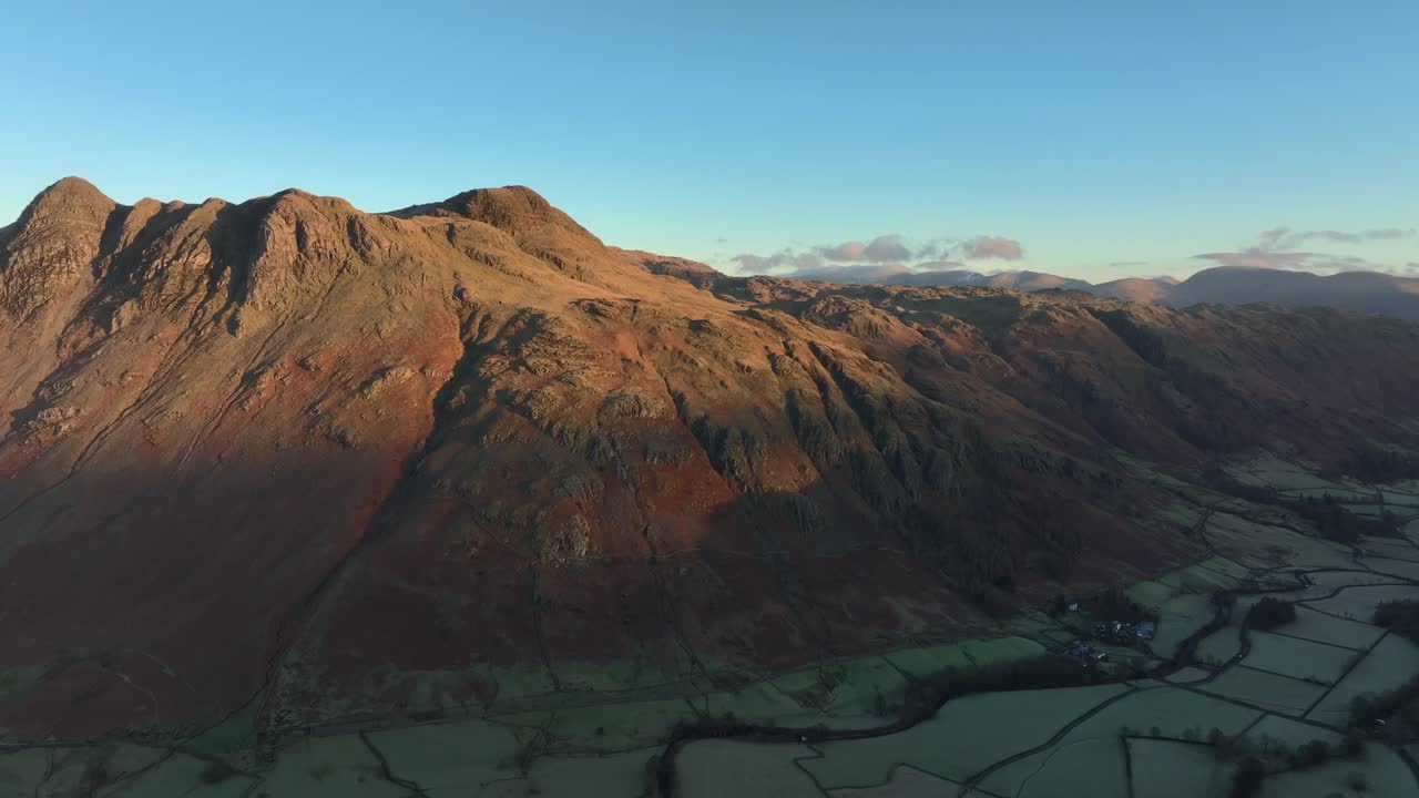 Flight over deep shadowed valley towards craggy mountains bathed in early morning wintry dawn light. Langdales, English Lake District, Cumbria, UK.