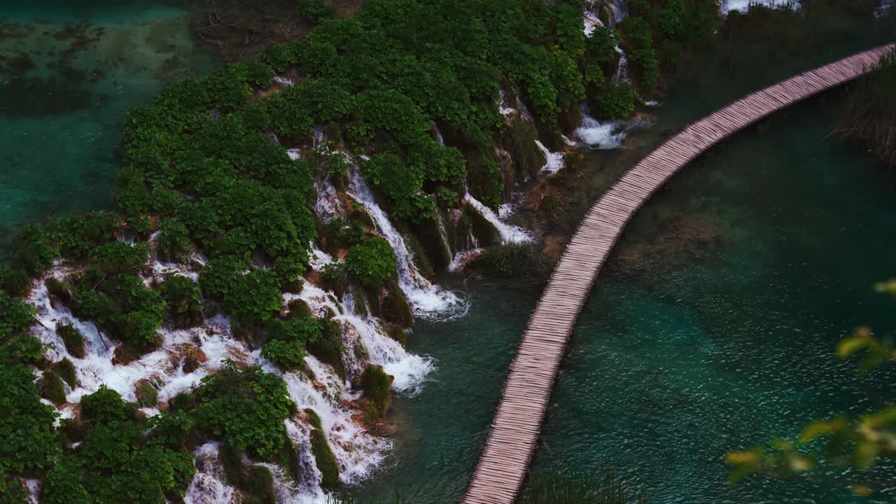 Waterfall flowing through mossy rocks into a clear lake with a wooden path in Plitvice National Park