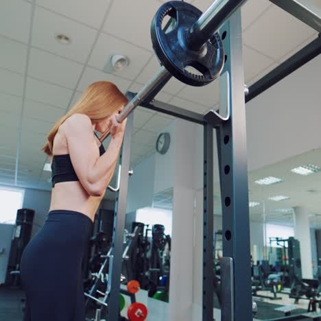 a young woman with a beautiful figure and flowing hair is doing an exercise with a barbell for arm muscles in front of a mirror in a gym.