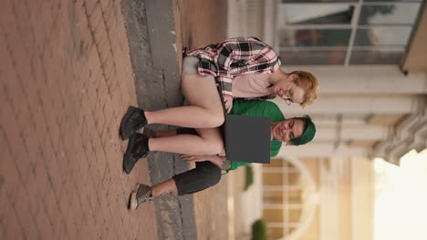 Vertical-video:-two-lesbian-girls-sit-on-the-steps-in-the-city-and-look-at-a-gray-monitor-screen-during-their-date.-A-blonde-girl-with-a-short-haircut-in-a-checkered-pink-shirt,-together-with-her-girlfriend-with-a-short-green-haircut-in-a-green-shirt,-are-talking-and-watching-something-on-the-monito