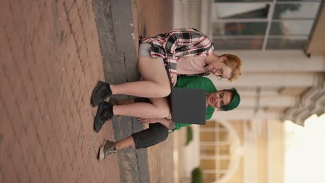 vertical video: two lesbian girls sit on the steps in the city and look at a gray monitor screen during their date. a blonde girl with a short haircut in a checkered pink shirt, together with her girlfriend with a short green haircut in a green shirt, are talking and watching something on the monito