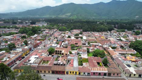 drone aerial footage of city block in antigua, guatemala colonial town showing bright and colorful red rooftops and green tree tops and forest mountain in the background