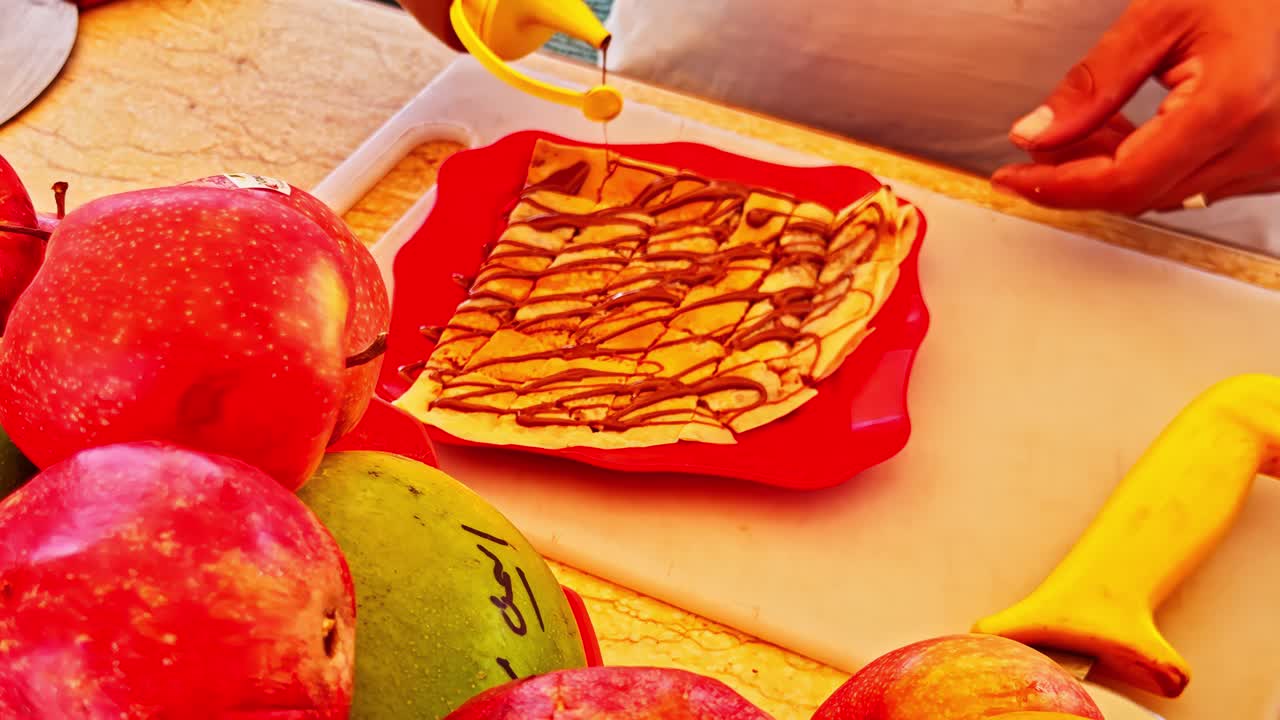 Street food vendor spreading chocolate sauce over Egyptian dessert at outdoor market stall, daytime scene