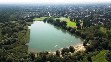 Explore-Un-Paisaje-Veraniego-Bañado-Por-El-Sol-Con-Un-Dron:-Un-Extenso-Campo,-Un-Lago-Sereno,-Bañistas,-Una-Animada-Cancha-De-Baloncesto-Y-Un-Animado-Campo-De-Fútbol.