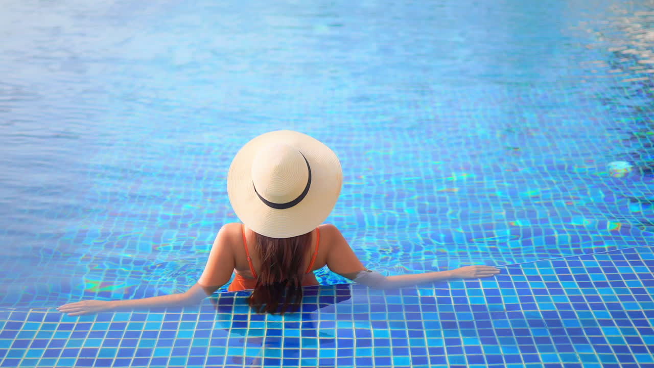With her back to the camera, a woman wearing a straw sunhat lounges in a swimming pool
