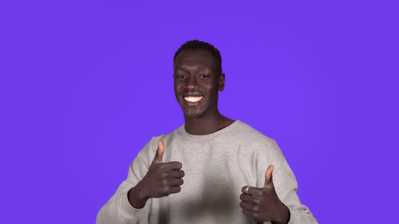 un joven afroamericano sonriendo y bailando de buen humor sobre un fondo azul. diversión imparable, felicidad, pulgares hacia arriba, emociones humanas