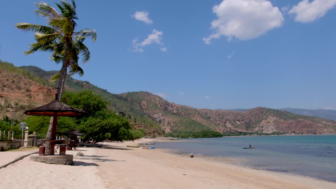 A scenic, remote white sandy beach, turquoise ocean water and Timorese fishermen in traditional fishing canoe on a windy day with coconut palm tree blowing on tropical island in Dili, Timor Leste