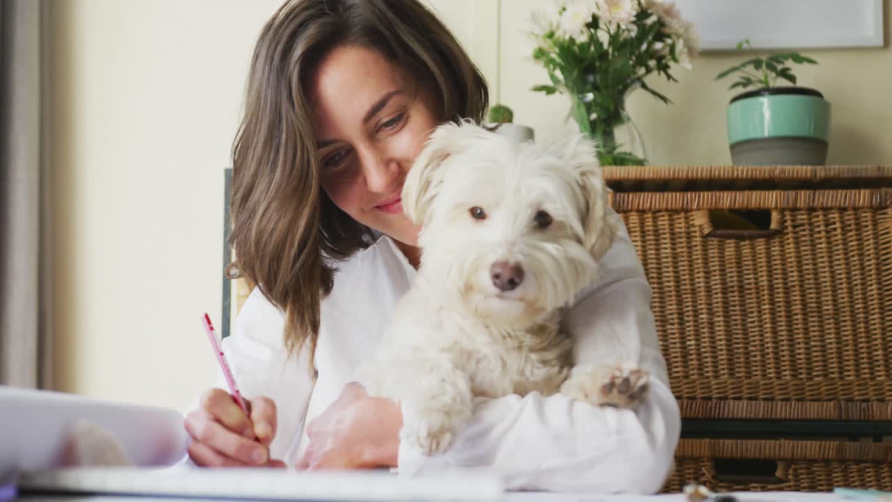 mujer caucásica dibujando trabajando desde casa y acurrucando a su perro de compañía