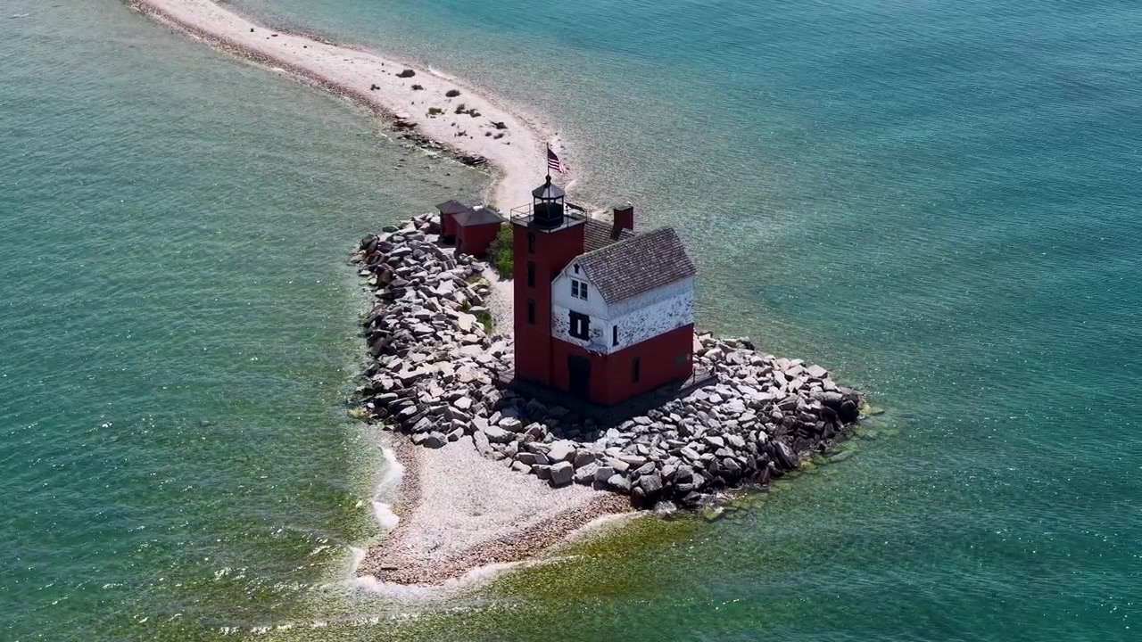 Vista obliqua con drone del faro di Round Island che si erge su una punta rocciosa accanto a una spiaggia di sabbia chiara nelle acque limpide dei Grandi Laghi.