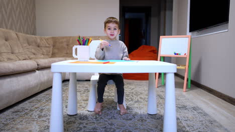 child creates at home. a young boy sits at a white table drawing and creating with colored pencils in a cozy living room setting