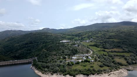 Aerial-drone-footage-of-small-cluster-of-buildings-nestled-by-a-river,-bridge,-and-dam