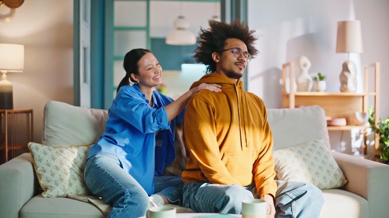 Asian girlfriend giving shoulder massage to boyfriend on sofa. Couple at home