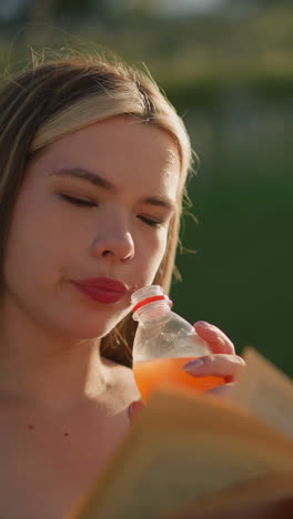 mujer sentada al aire libre con un libro en una mano y bebiendo jugo de una botella en la otra, continúa leyendo, con un fondo borroso de vegetación y poste de lámpara