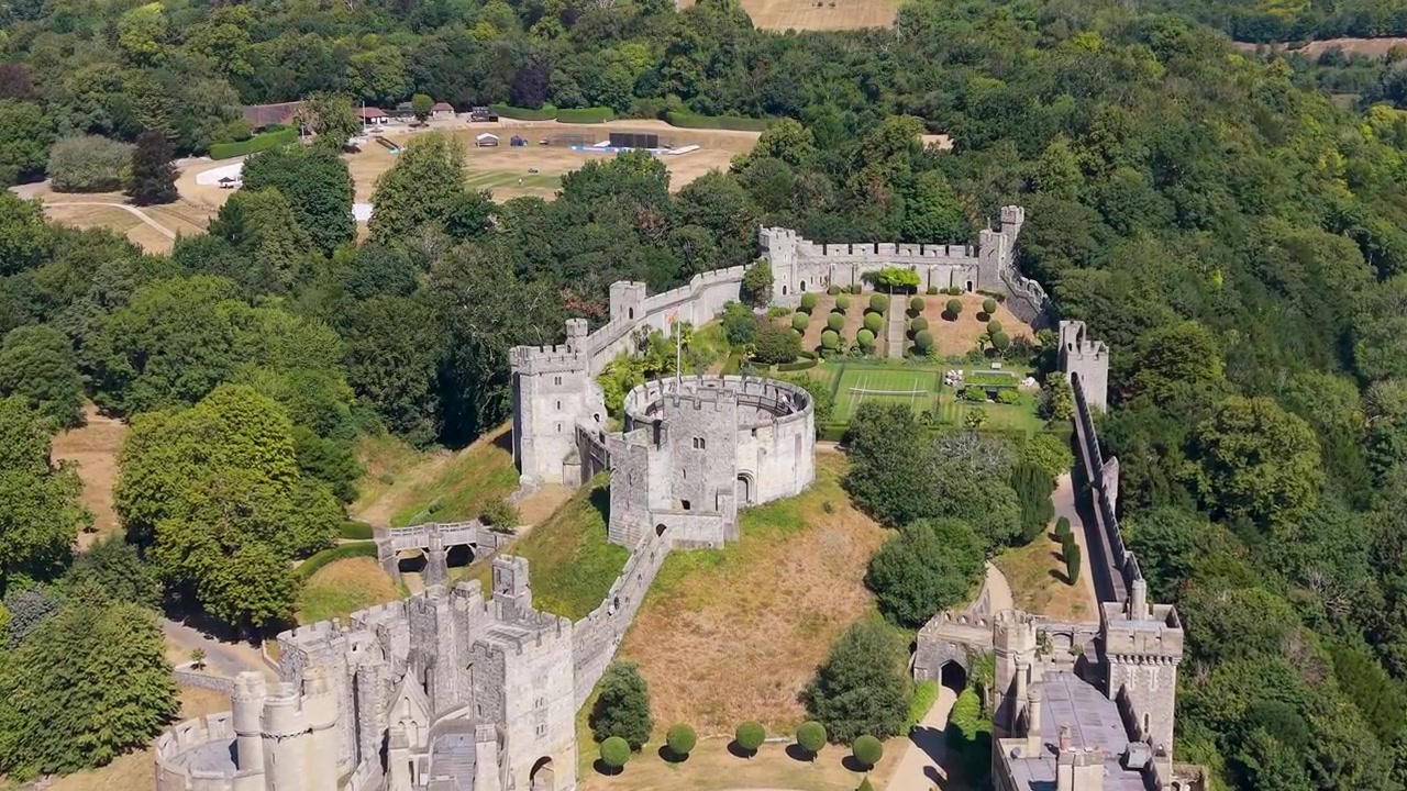 Survol aérien du château d'Arundel pendant une canicule estivale, révélant des pelouses jaunies par la sécheresse, des murs et tours en pierre historiques, et une dense canopée d'arbres entourant le monument dans le West Sussex, Angleterre