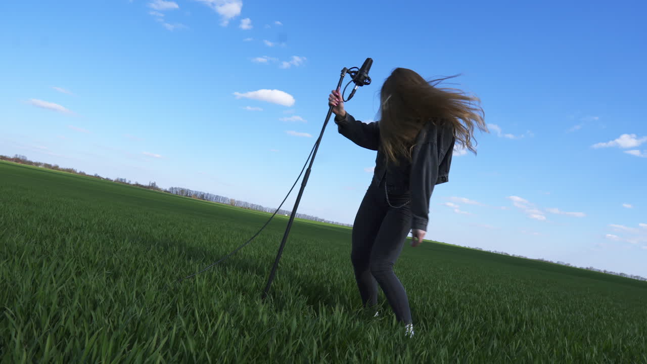 Cheerful happy young lady dancing emotionally near the microphone stand in the green field. Female having fun while making video for music clip.