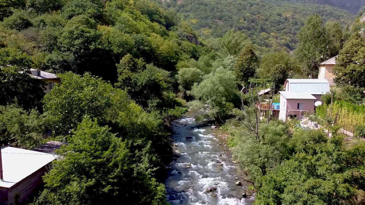 A river gently flows through a picturesque village surrounded by rolling hills and dense trees. The tranquil scenery invites relaxation and connection with nature