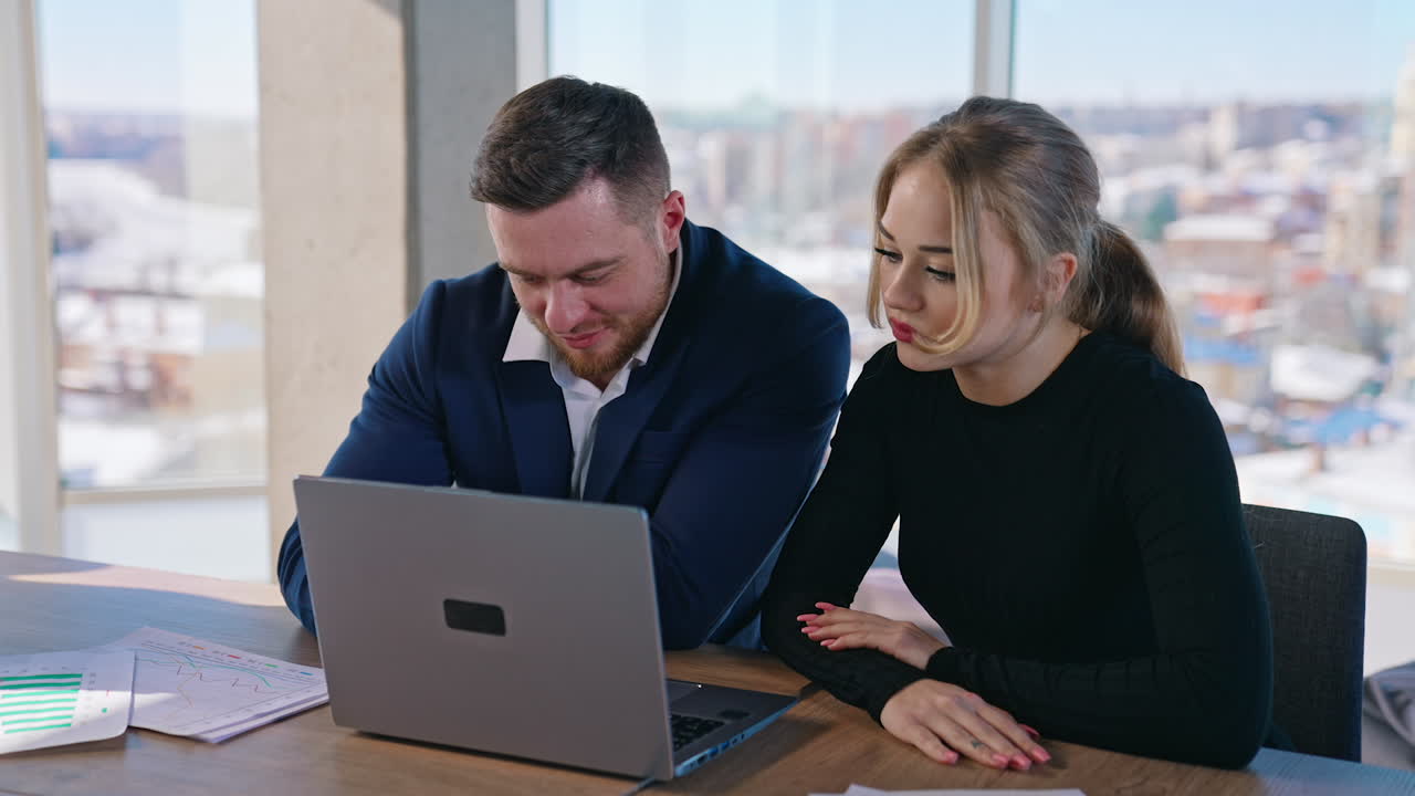 Young pretty collegues work on business. Beautiful female and young businessman sitting together at the table and look into a laptop in office room on city window background.