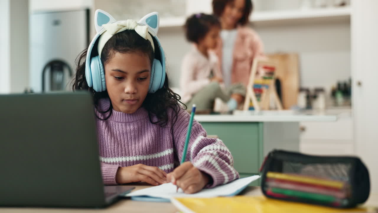 Young girl working on her laptop at home, with her mother and sibling playing in the background