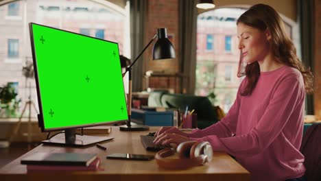 joven mujer adulta hermosa trabajando desde casa en una computadora de escritorio con pantalla verde. mujer creativa comprobando y escribiendo correos electrónicos. apartamento loft con vista a la ciudad urbana desde una gran ventana.