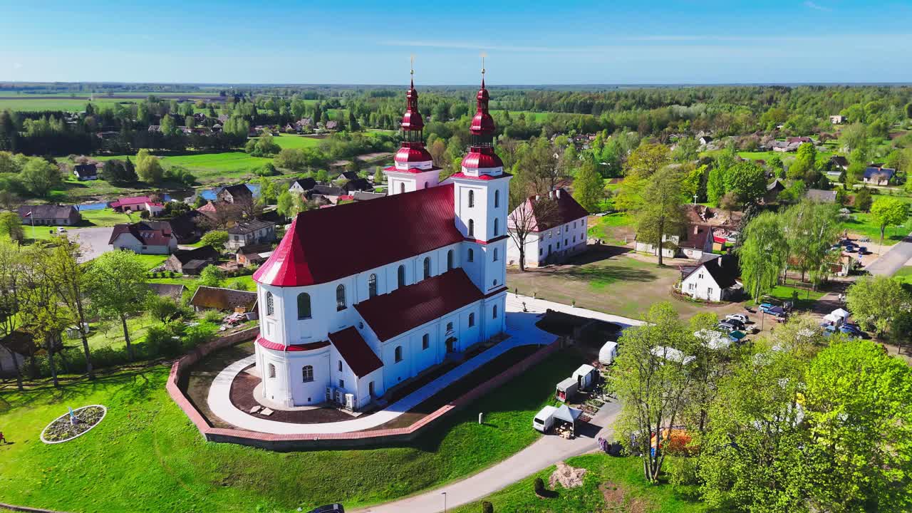 A white church with twin red spires rises from a serene European village landscape, framed by cottages, trees, and open farmland in clear, sunny weather.