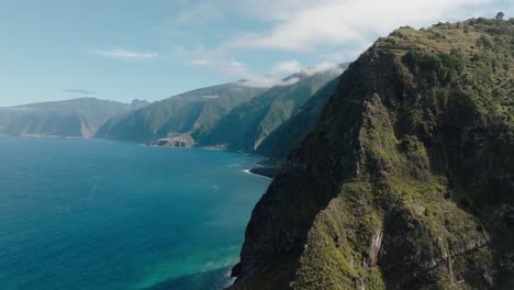 sweeping aerial of madeira’s northern cliffs near ilhéus da ribeira da janela, revealing winding roads, green peaks, and deep blue atlantic waters.