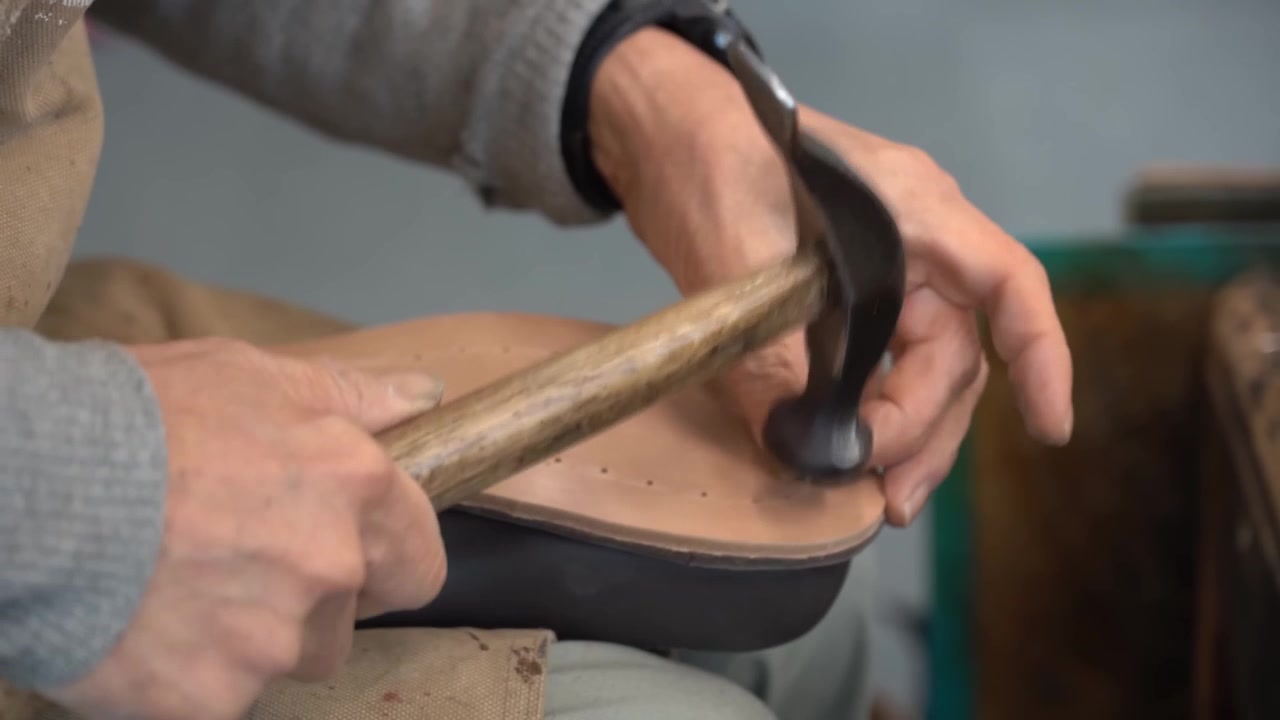Craftsman tapping a hammer along a tan leather midsole punched with stitch holes while it rests on a wooden last