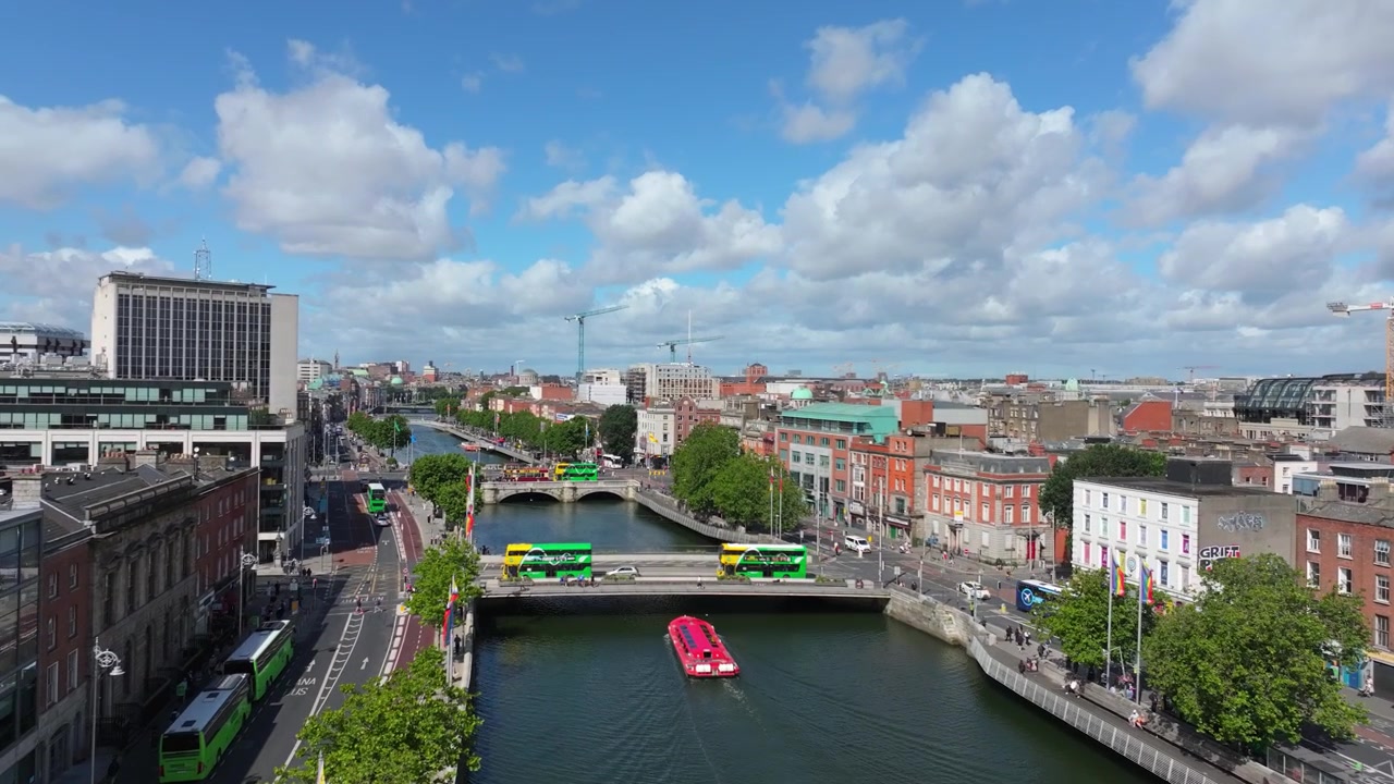 Der Fluss Liffey fließt durch das Zentrum Dublins, während ein leuchtend rosa Ausflugsboot Brücken und Stadtverkehr unter blauem Himmel passiert