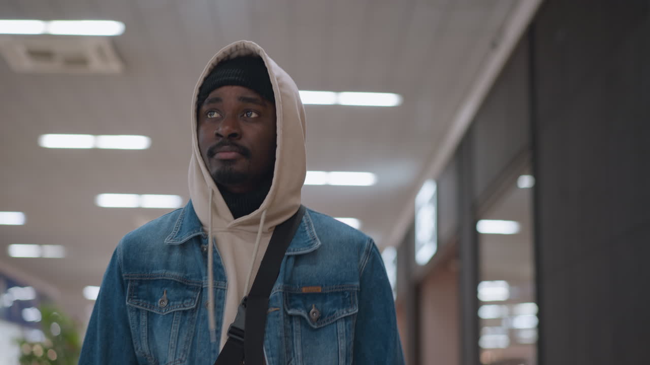 Close up view of man wearing denim jacket layered over hoodie and beanie cap looking upward in modern mall corridor while carrying bag strap visible against illuminated ceiling panels and blurred