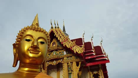 dolly out shot of a golden buddha statue and thai buddhist temple with ornate style of high gables, stepped-out roofs and elaborate finials in an overcast weather in thailand asia