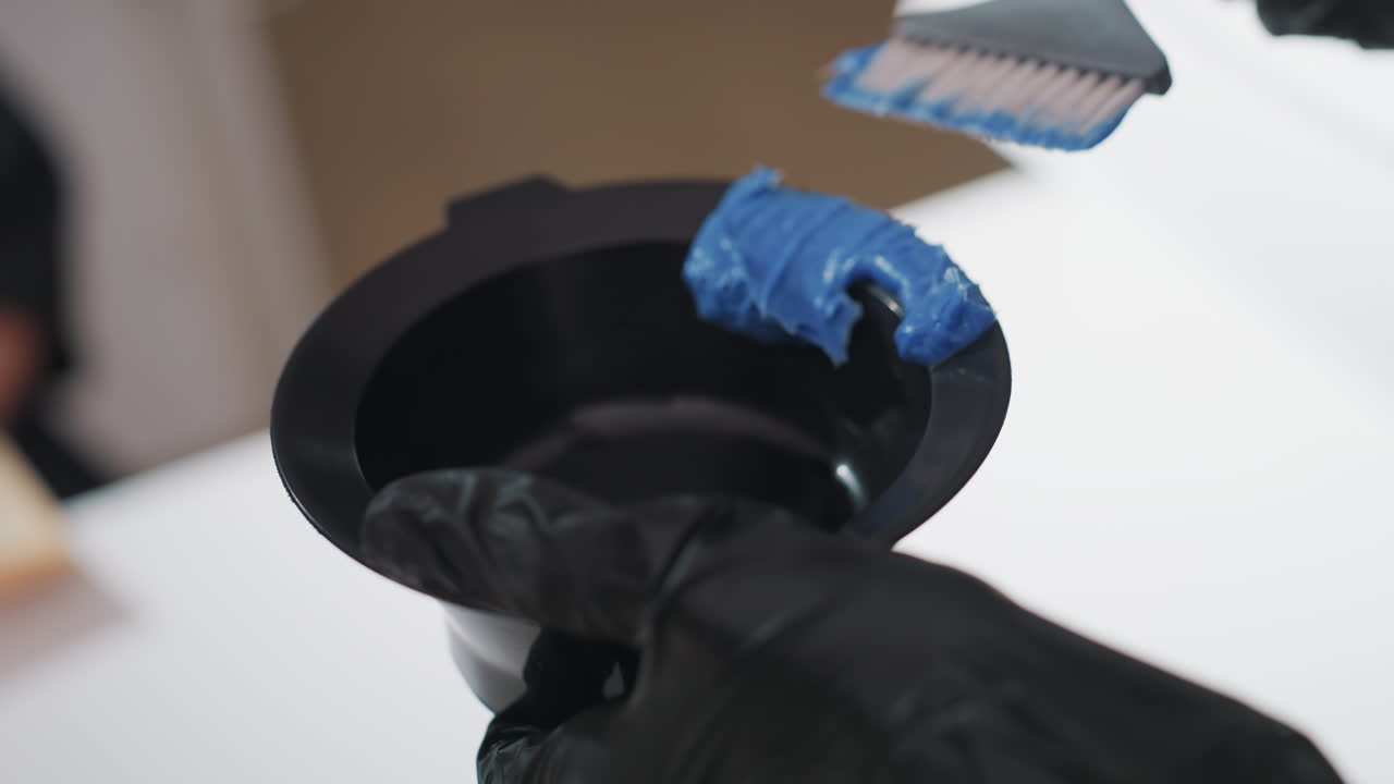 ayudante con guantes vertiendo tinte azul en un cuenco negro, transferencia cuidadosa del pincel al recipiente, superficie blanca brillante y manos firmes, preparación en el salón antes de la aplicación al cabello del cliente