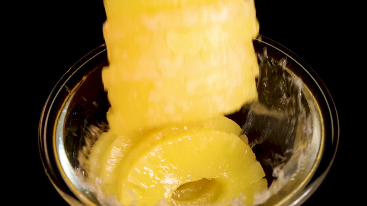 Stacked pineapple rings fall into a clear glass bowl, creating a dramatic splash. High-speed close-up, bright lighting, black background, and static camera