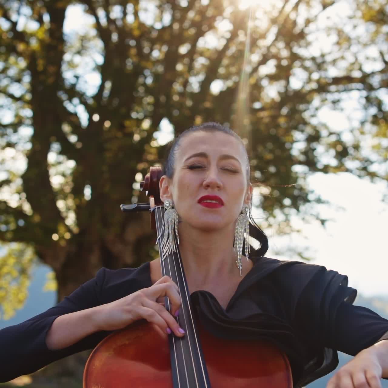 Soulful profound music performance by a female musician. Woman in black dress and long earrings playing bass-viol at the backdrop of beautiful summer nature