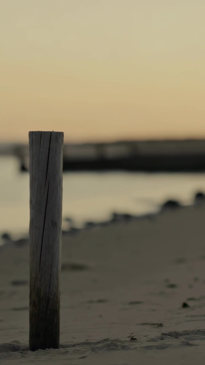 Vídeo vertical: Câmera se movendo pela areia, acompanhando um poste que entra pela esquerda, centralizando o poste com um píer distante.