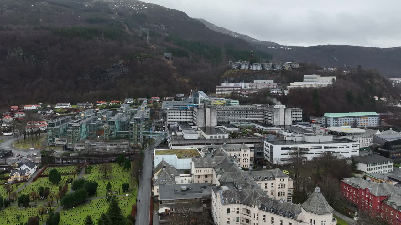 Haukeland Hospital and Helse Bergen in Norway with a graveyard on the side and mountain backdrop. Slow aerial rise on an overcast day