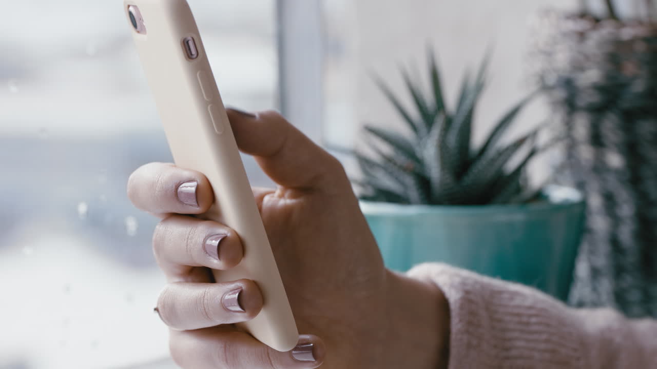 mano de mujer de cerca usando un teléfono inteligente navegando por mensajes en línea leyendo redes sociales disfrutando de la comunicación móvil de pie junto a la ventana relajándose en casa en un día frío y lluvioso