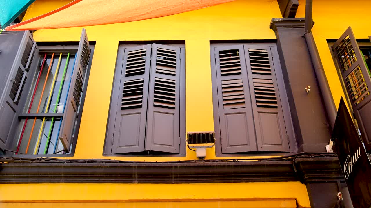 A brown wooden shuttered window on a vibrant yellow wall is gradually opened, revealing architectural details. Daylight, static camera, upward angle
