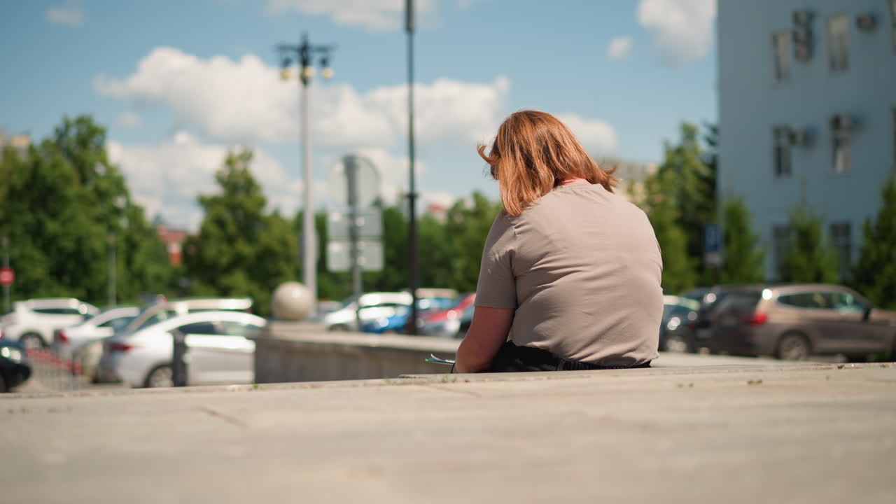 Vista traseira de uma mulher sentada ao ar livre, desenhando em uma prancheta sob a luz do sol, com o cabelo suavemente movido pela brisa, cercada por paisagens urbanas com árvores verdes e carros