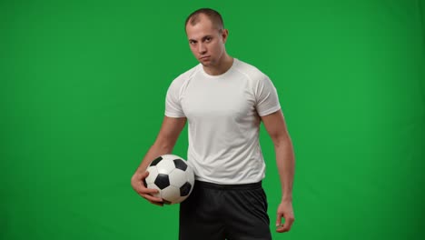 joven deportista enfocado atrapando una pelota de fútbol con la pierna en el fondo de chromakey y mirando a la cámara. retrato de un futbolista caucásico profesional serio posando en cámara lenta en la pantalla verde.