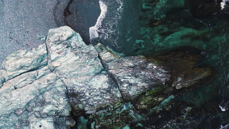 ocean waves crash on grey shoreline with white rocky outcropping, drone top down