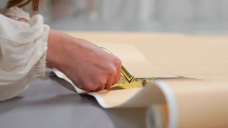 Young female designer with tape-line on her neck standing in dressmaking studio and drawing lines with chalk and rule. Female couturier in atelier cutting out a pattern for future clothes.