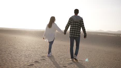 Rare-view-of-couple-holding-hands-walking-in-romantic-relationship-under-sun-and-blue-sky-in-desert.-Two-young-lovers-walking-by-desert-sand-in-casual-clothes,-holding-hands.-Lens-flares