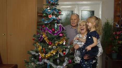 Senior-grandfather,-grandmother-and-granddaughter-child-lights-garlands-on-artificial-Christmas-tree