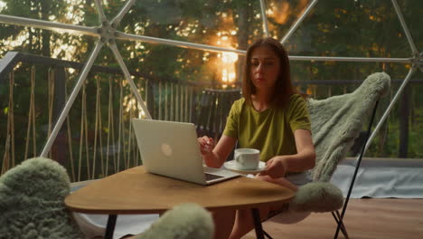 busy woman drinks tea in cup working in online sphere in tourist house surrounded by forest. business trip to another country with incredible views at sunset slow motion