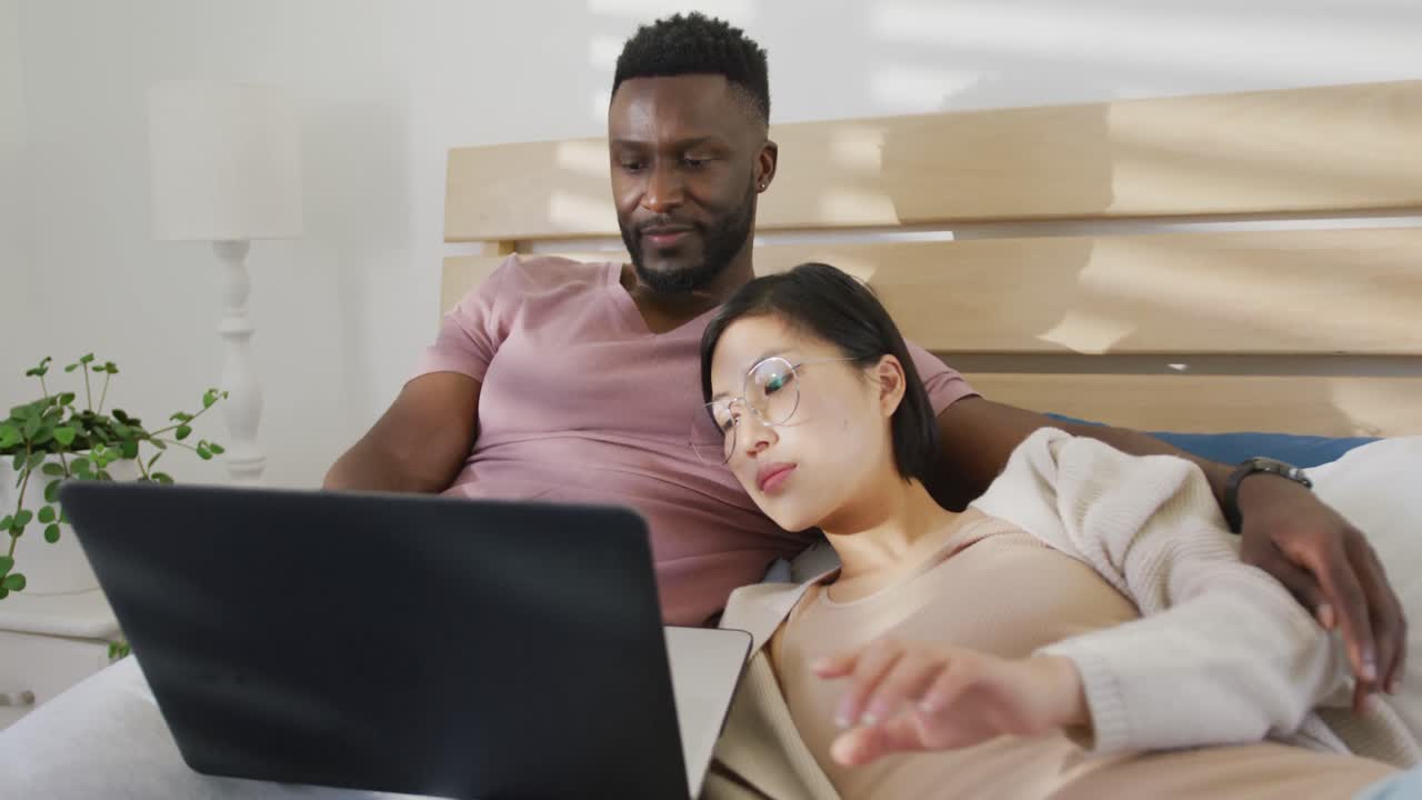 Happy diverse couple using laptop and lying in bedroom